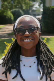 A smiling female student with glasses, white shirt, long dark hair in intricate braids and headband