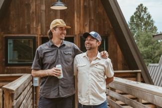 Two men wearing baseball caps stand arm-in-arm smiling on a wooden deck in front of an A-frame cabin.