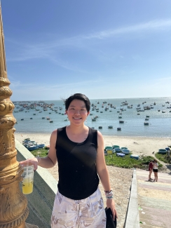 A smiling person with short dark hair and a black tank top holds a cold drink while standing on stairs overlooking a sandy beach dotted with round coracle boats and floating vessels on clear blue ocean water.