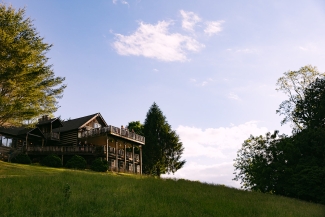A large wooden log lodge with multi-level outdoor balconies stands atop a steep grassy hill under a blue sky.