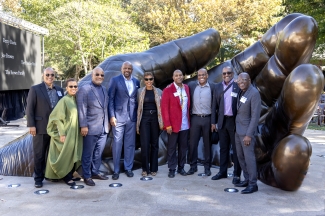 A group of eight people poses smiling outdoors in front of a large bronze sculpture of open hands.