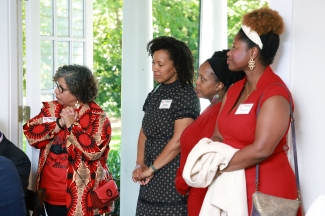 Four women stand in a line indoor near a doorway, listening attentively to a presentation.