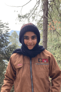 A female student in a forest with a hat and scarf covering her hair, brown canvas jacket, and a mountain view in the background