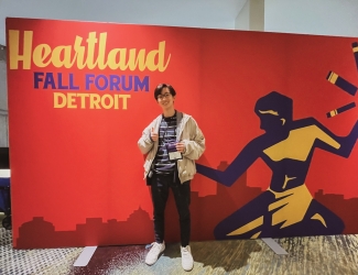 A young man smiling and giving a thumbs-up standing in front of a red "Heartland Fall Forum Detroit" backdrop.