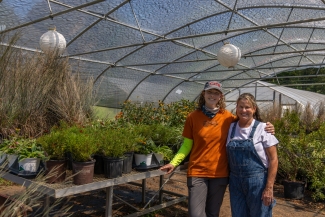 Two women in outdoor work clothes in a greenhouse, filled with native plants in pots