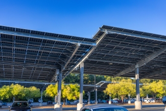 A large solar panel canopy structure shades a parked car lot under a clear blue sky, with trees visible in the background.