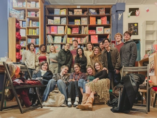 A diverse group of smiling young adults poses together inside a cozy bookstore with tall shelves filled with children's books and illustrations in the background.