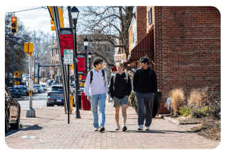 Three young men wearing backpacks walk and talk together down a brick sidewalk lined with brick buildings and streetlights.