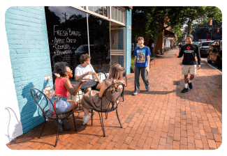 A group of young adults socializes outside a café on a brick sidewalk next to a turquoise wall with a handwritten menu.