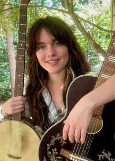 a smiling young woman with long brown hair, holding a banjo and a guitar in a forest