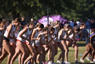 A line of female cross-country runners in matching red and white uniforms crouch at the starting line on a grassy field.
