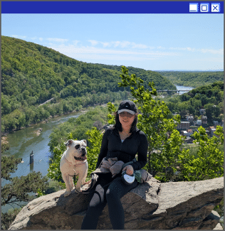 A woman on a hike with her dog, both seated on a rock above a gorge full of trees.