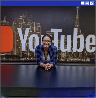 A woman with her clasped hands on a desk in front of the YouTube logo and a skyline