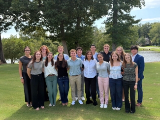 A group of young adults in business casual attire pose together on a grassy lawn with trees and a body of water in the background.