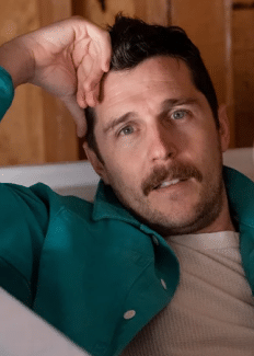 a man in a bathtub, brown hair and mustache, resting his head in his hand