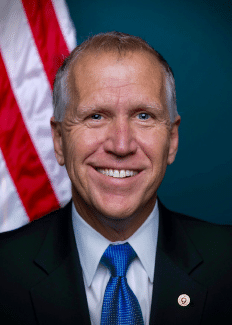 Representative Tom Tillis with a blue tie, black suit in front of a US flag