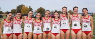 Eight female cross-country runners wearing matching red and white Davidson College uniforms and race bibs pose together in a line outdoors.