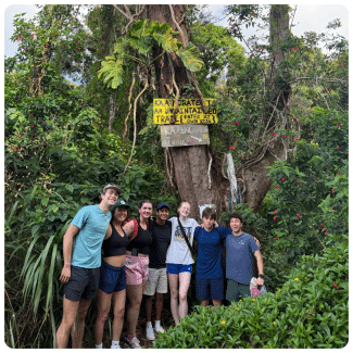 Seven hikers pose together in dense tropical forest under a yellow warning sign reading Ka'au Crater Trail is an unmaintained trail enter at own risk.