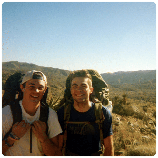 Two young men wearing large backpacks smiling together during a hike through a sunny desert landscape.