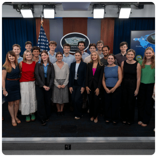 A group of young adults poses for a group photograph inside the Pentagon press briefing room, standing in front of an American flag and the Pentagon seal.