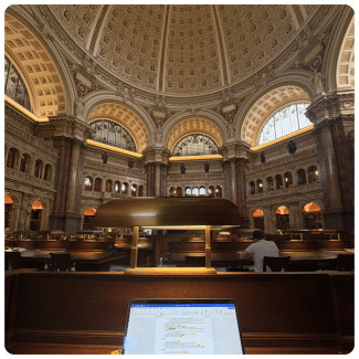 A view from a study desk with an open laptop and brass lamp looking out into the grand Main Reading Room of the Library of Congress, featuring ornate arched windows, marble columns, and a large coffered dome.