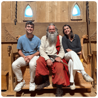Alt Text: A young man and woman sit smiling on an ornate wooden bench on either side of a man costumed as a Viking reenactor with a long grey beard and historical garments.
