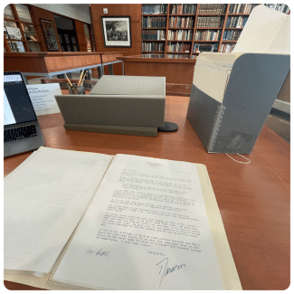 An open archival folder containing typed letters rests on a library research desk beside a laptop, book cradle, pencil cup, and document storage box.