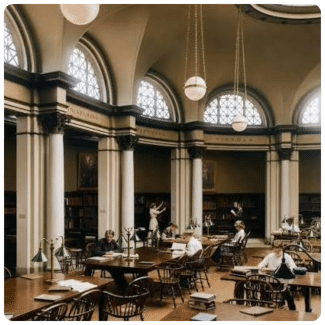 People study at wooden tables inside a grand, classical library reading room featuring arched windows, round pendant lights, and tall columns.