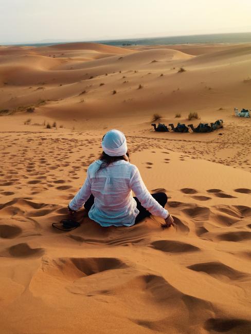 Student sits on sand hill surrounded by desert facing away from the camera.