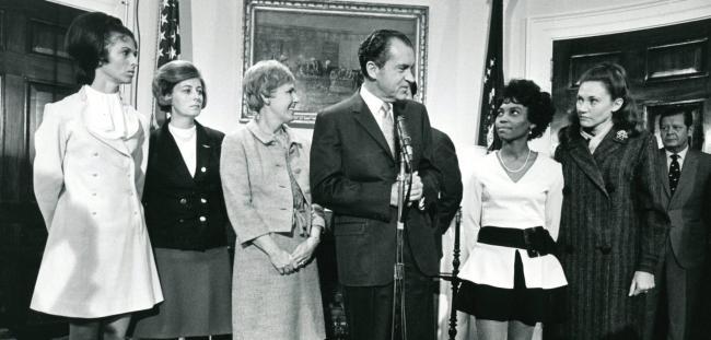 Sybil Stockdale to the left of President Nixon speaking at microphone surrounded by other women in the National League of Families