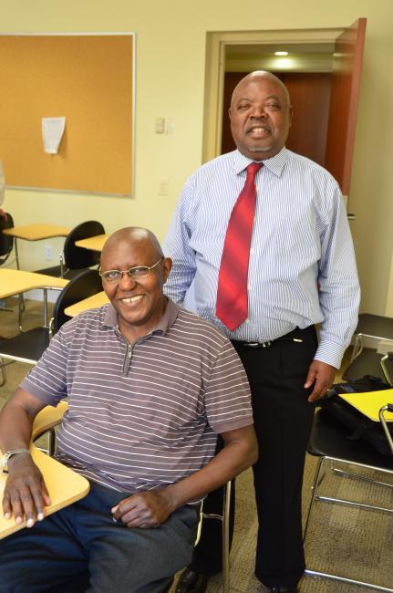 Grown-up Georges Nzongola-Ntalaja '67 standing and Benoit Nzengu ’66 seated in a desk in a Chambers building classroom at Davidson
