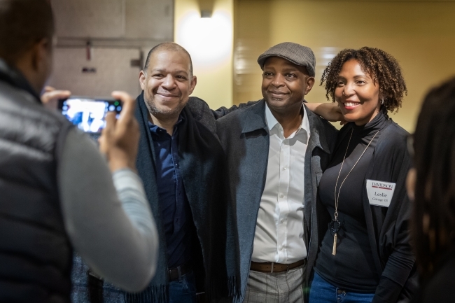 two men and a woman smiling as someone in foreground takes their picture