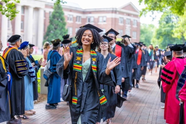 Student smiles and waves while wearing graduation regalia