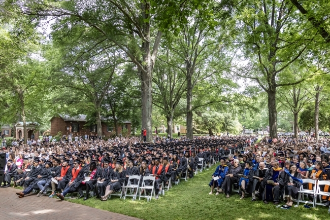 Lawn of chairs and people seated in them outdoors under a canopy of trees