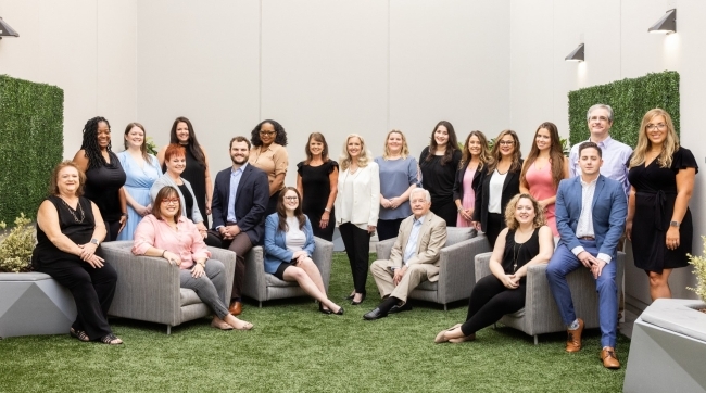 A group of 30 lawyers sit together on grey couches with green walls in the background.