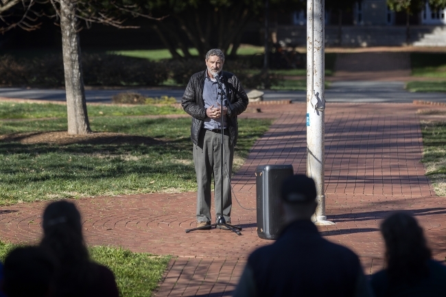 A white man wears a flannel shirt and jacket and speaks to a crowd of people outdoors