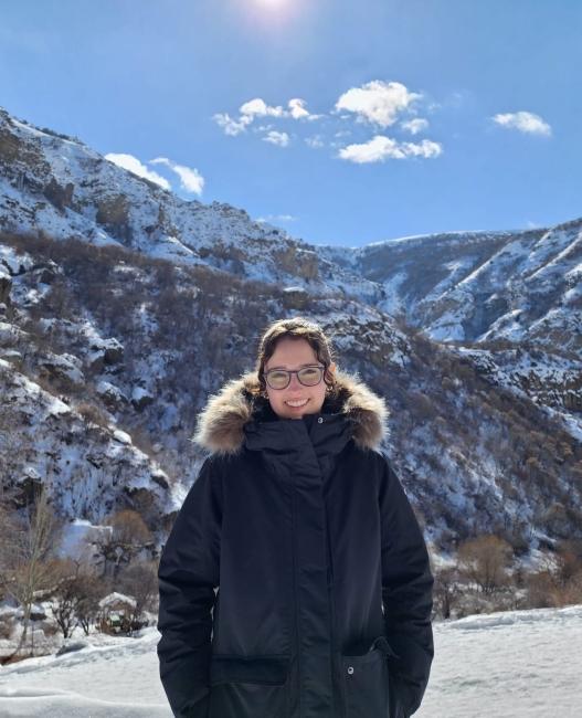 A white woman wears glasses and a big winter coat, stands smiling in front of mountains