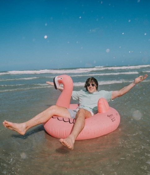 A young white male sits in a flamingo inflatable tube on a beach