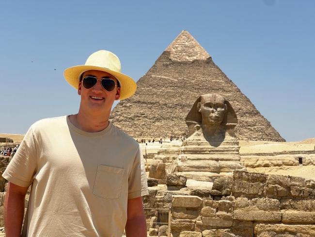 a white male wearing a tshirt and straw hat smiles in front of a pyramid with blue skies in the background
