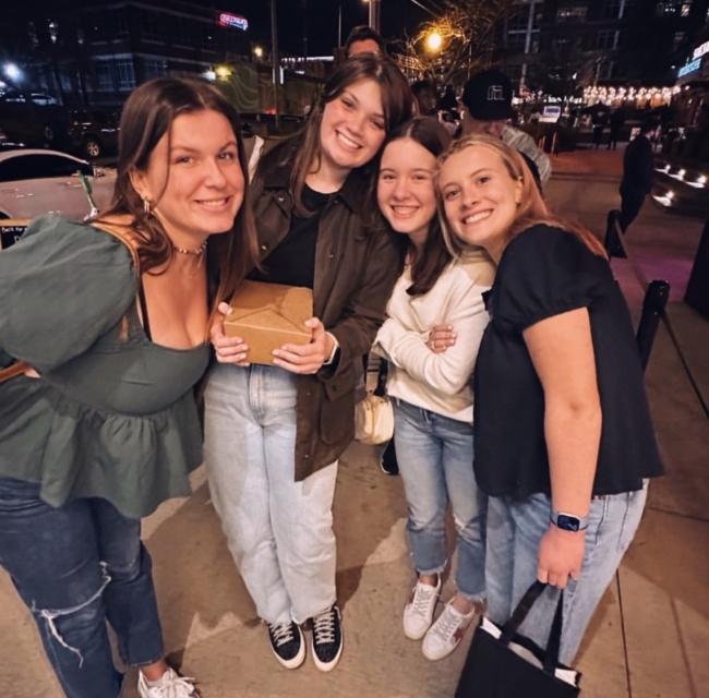 a group of young women wearing jeans and tops smile while standing on a city street at night