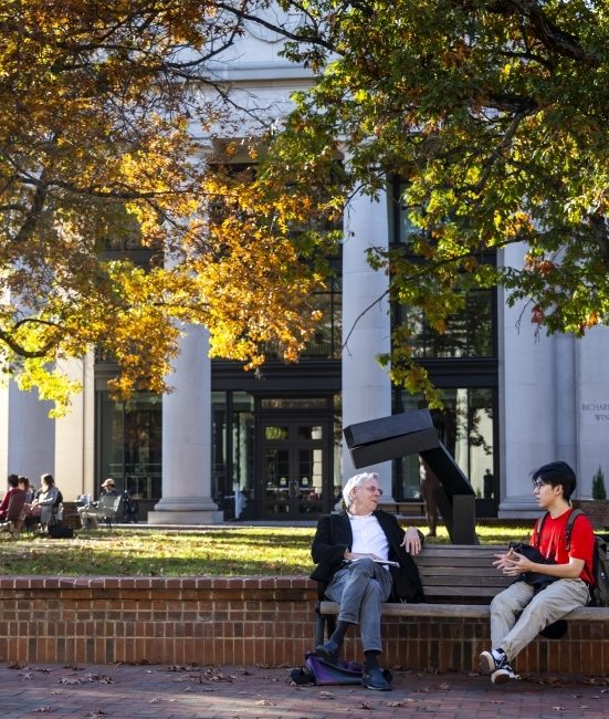 a faculty member and a student sit on a bench outside and have a conversation