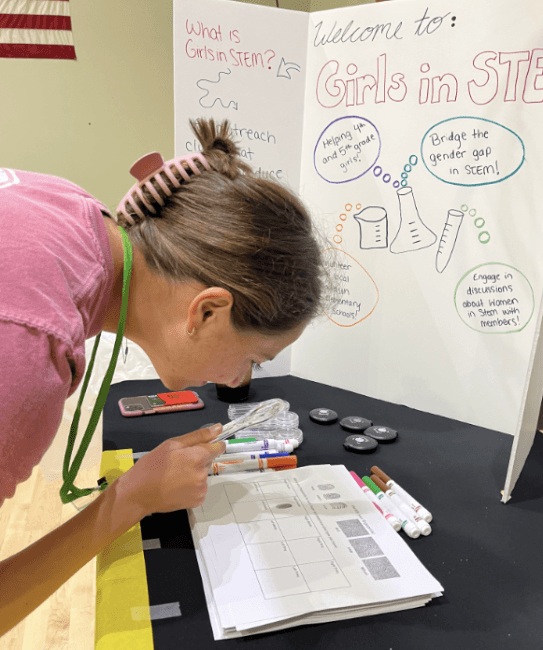 a young woman holds a magnifying glass while standing over a table with a sign "Girls in STEM" in background