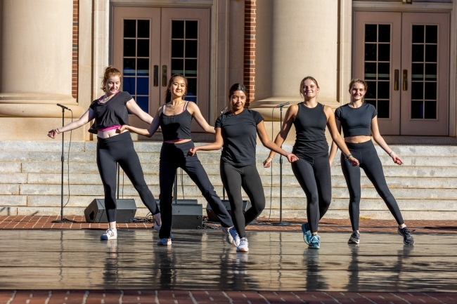 a group of five students in all black outfits dancing and singing on a stage