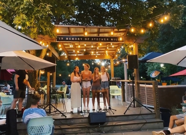 a group of young women sing on a stage outside as twinkle lights are overhead