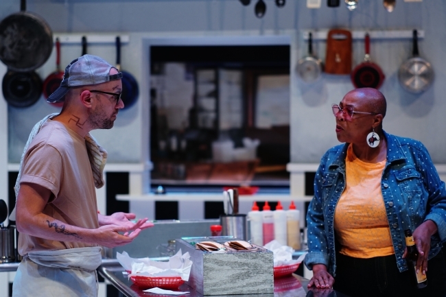 on a stage, a white person argues with a Black person while standing around a restaurant kitchen table