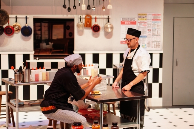 on a stage in a restaurant kitchen, a Black man sits at a table while talking to a white man standing