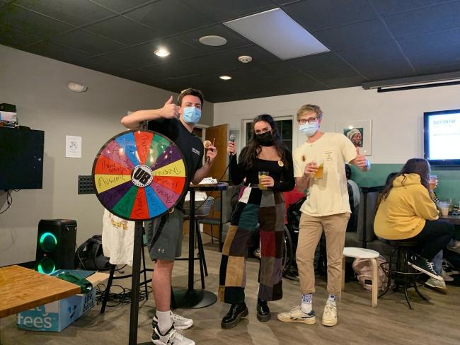 a group of students in masks stand around a wheel and hold thumbs up