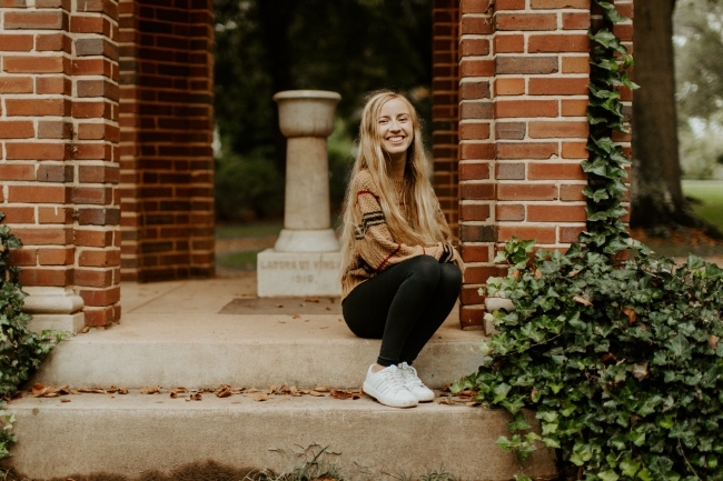 a young white woman wearing black leggings and a sweater sits on a step outside and smiles