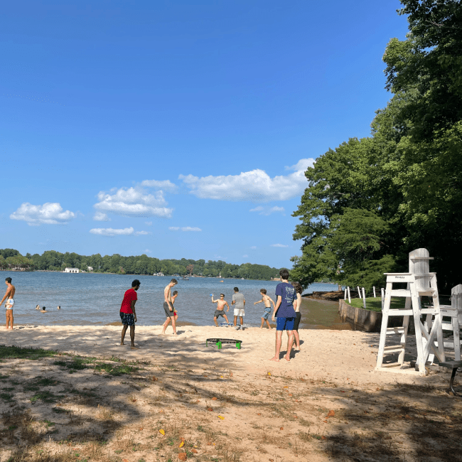 a group of students stand around a beach and swim in a lake on a sunny day