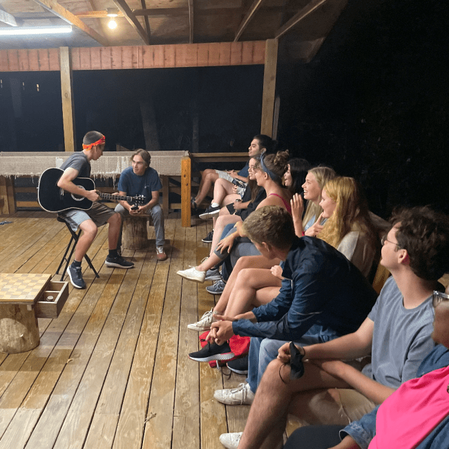 a group of students sit around a cabin as one plays the guitar